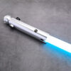 Close-up of the highly detailed silver Snips CW primary saber hilt, featuring a detailed control box with red, black, and toggle switches, resting on a textured concrete surface with the blue electronic blade glowing.